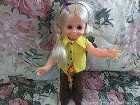 Front view of vintage blonde Chrissy doll named Velvet seated on floral fabric. Dressed in yellow top and brown pants with hair tied with purple ribbon.
