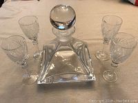 Photo showing one large square-based crystal decanter surrounded by four sherry or liqueur glasses on a flat surface.