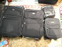 Five black luggage pieces: three upright suitcases of different sizes, one backpack, and one suit bag, all arranged on floor.