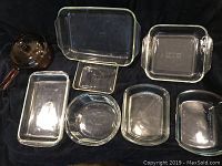 Nine Pyrex glass baking dishes of various shapes and sizes including a covered saucepan shown on a dark cloth background.