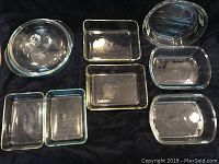 Nine clear glass baking dishes arranged on black fabric. Includes a round covered casserole, six small rectangular dishes, and one oval dish.