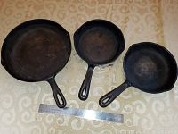Top-down view of three cast iron skillets of different sizes arranged side-by-side with a measuring ruler below for scale. All skillets have long handles with hanging holes and seasoned black cast iron surfaces.
