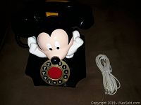 Black rotary dial phone with large Mickey Mouse figure holding handset, and white telephone cord.
