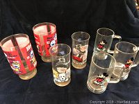 Photo shows seven vintage glasses including Hee Haw tall glasses, ET character glasses, and two Mickey Mouse mugs with handles on a black fabric background.