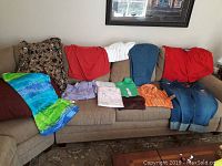 Photo showing varied women's clothing laid out on a beige couch, including tie-dye dress, floral blouse, solid color tops, denim pants.