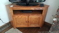 Front view of wood grain corner console cabinet with television on top, showing two cabinet doors and open shelf.