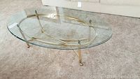 Oval glass top brass coffee table showing the clear glass and brass frame with clasp attachments and carpeted floor.
