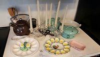 Wide view showing enamelware pots, deviled egg plates, candelabras with candles, teapot, utensils, pink bisque soap dishes, and iron trivet on counter