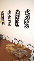 Photo showing four vertical cast metal wall art pieces with detailed leaf designs mounted on white wall above a table with other decorative items.