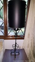 Front angle showing one black metal table lamp with a tall cylindrical shade on a wooden table near a window.