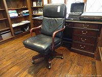 Full front and side views of black leather-feel office chair with carved wooden arms and wooden base with casters on hardwood floor in office setting.