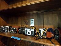 Shelf holding five 35mm film cameras, four camera cases, and one compact exposure meter.