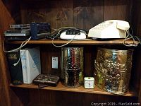 Shelf with two vintage tape decks, two telephones (white rotary and black push-button), two metal relief embossed trash bins, two hardcover books, and a vintage wind-up alarm clock.