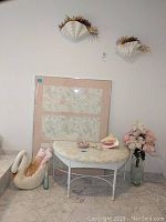 Bench with upholstery, framed art behind, seashells and shell soaps on bench, decorative ceramic swan container, glass vase with flowers.