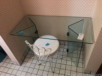 Photo showing glass vanity with glass top and glass panel supports, alongside metal chair with white cushioned seat in tiled room corner.