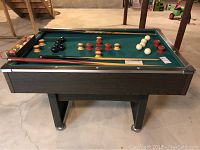Full view of bumper pool table with balls and cues placed on the table in a basement setting with concrete floor.