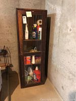 Front angled view of vintage wooden cabinet with glass door, showing stored cleaning supplies on shelves inside. Visible wear such as scratches and finish loss on the wood frame, placed in a basement with concrete walls.