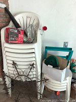 Stack of white plastic chairs and garden accessories including hose inside a box, battery charger, and leaf blower nozzle visible on chair top