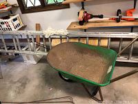 Photo showing green wheelbarrow and aluminum roofing ladder side by side in garage environment.