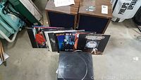 Photo showing two wooden speakers with blue fabric fronts, a pile of classic rock vinyl albums, and black turntable with dust cover on a concrete floor.