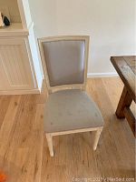 Front view of one of four light beige upholstered dining chairs with square backs on a wooden floor near a dining table.