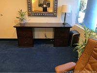 Front view showing dark wood desk with two drawer pedestals, metal handles, lamp and plant on top, mounted under a decorative mirror.