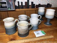 Four tea cups, one creamer, and two tall goblet-style cups with speckled white to blue glaze on wooden shelf.