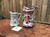 Two china creamers or pots displayed outdoors on a wooden table. Larger creamer on the right with ornate floral design and handle. Smaller on the left with green rim and fruit/flower motifs.
