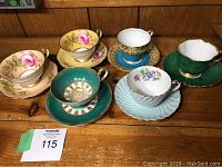 Six assorted English bone china tea cups and matching saucers with floral and gold designs on display on wooden surface.