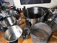 Group photo showing all cookware items together including pots, steamer, frying pan, mesh basket, and lids, on wooden surface.