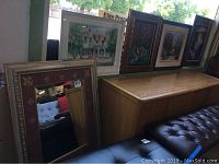 Seven pieces of wall art and a large mirror arranged near a wooden cabinet and black tufted bench. Includes a large mirror with purple and gold fleur-de-lis border and framed paintings depicting houses and other themes.