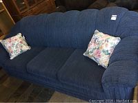 Front and right side view of a blue fabric sofa with floral cushions in a room with stone flooring and other furniture items visible around.