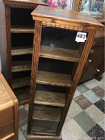 Two medium oak finished wooden swivel corner cabinets shown side by side, each with glass front door and four shelves inside. Cabinets have grooved decorative edges.