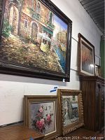 Wall of five framed artworks including large oil painting of cafe by canal, floral still-life, and architectural scenes.