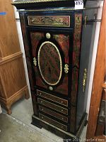 Front view showing full height of Asian inspired cabinet with intricate red and gold painted wood and metal accents. Includes two rows of drawers and large door with oval design.