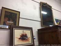 View of multiple wall art pieces including framed paintings and framed vertical mirror stacked against wall