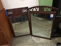 Photo showing two wall mirrors with dark wooden frames featuring an arched top geometric cutout design, placed on concrete floor leaning against a wall.