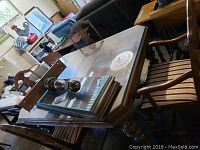 Dining table with glass top and extra leaves stacked under it, showing wood base and one armchair in striped upholstery