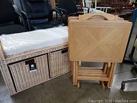 Wicker storage bench with white cushion on top next to folded wooden TV trays in front of black office chairs, showing front and side view