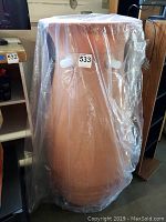 Image shows a tall terracotta colored plastic rain barrel wrapped in clear plastic with two white spouts visible.