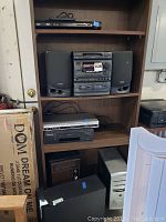 Full view of shelving unit holding various electronics including Aiwa audio system with speakers, Toshiba DVD player, Mitsubishi cassette player, and external hard drives.