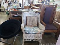Set of three chairs including white wicker rocker with cushion, tall wooden framed upholstered chair, and small black circular lounge chair with metal frame.