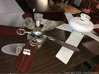 Two ceiling fans displayed on a table; one fan with dark wooden blades and five light fixtures, the other with white blades and a central perforated light fixture. A remote control is visible near the dark wood blade fan.