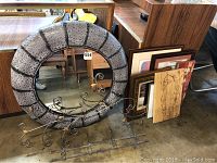 Round framed mirror with textured gray and black segmented frame leaning against wood furniture, accompanied by metal decorative wall hangers and framed artworks stacked nearby.