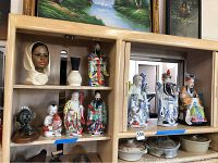 Wide view of 10 ceramic figurines arranged on wooden shelves: includes African style bust, white vase, colorful Asian figures in robes, and smaller figurines.
