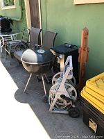 Photo shows portable Weber charcoal grill, Masterbuilt electric smoker, hose reel cart with garden hose, and a freestanding wooden post all arranged against a green wall.