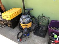 Photo showing stacked yellow and black plastic storage bins, large ShopVac Ultra Pro vacuum, smaller ShopVac vacuum, and small metal wire animal cage.
