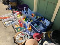Wide view of multiple plastic bins filled with an assortment of hand tools, cleaning supplies, hoses, and related items arranged on a patio surface.