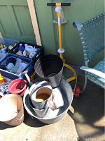 Full view of the yellow wheeled drum caddy leaning against a wall next to a gray metal tub, several plastic pots including black ribbed and beige ones, and a red small pot. The items are placed on a concrete surface.