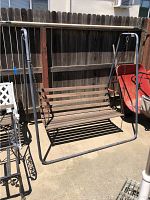 Photo showing the full freestanding metal porch swing frame with wooden slatted swinging seat in an outdoor setting, next to a wooden fence and some garden tools.